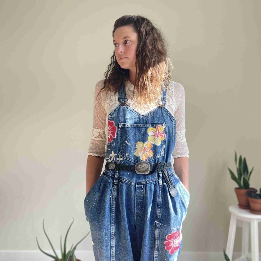 a woman standing in a room wearing a jean overall.