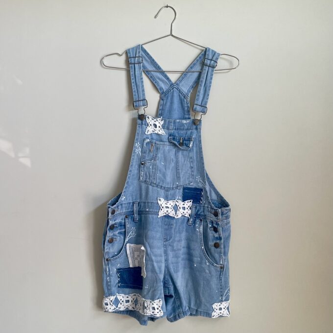 A Patchwork Upcycled Denim Short Romper Overalls hanging on a wall.