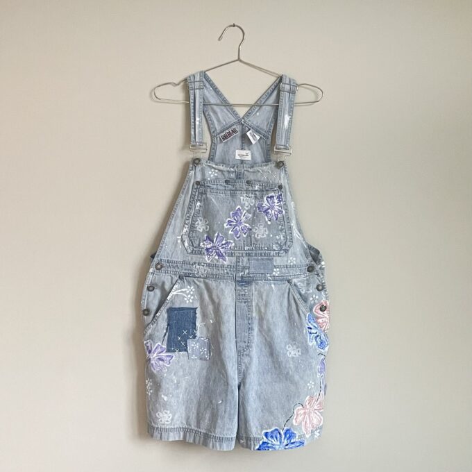 A pair of Painted Floral Patchwork Denim Romper Overalls hanging on a wall.