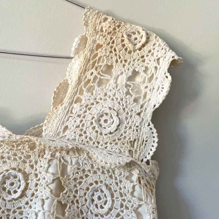 A tea stained cream crochet festival tank top with tie back hanging on a wall.