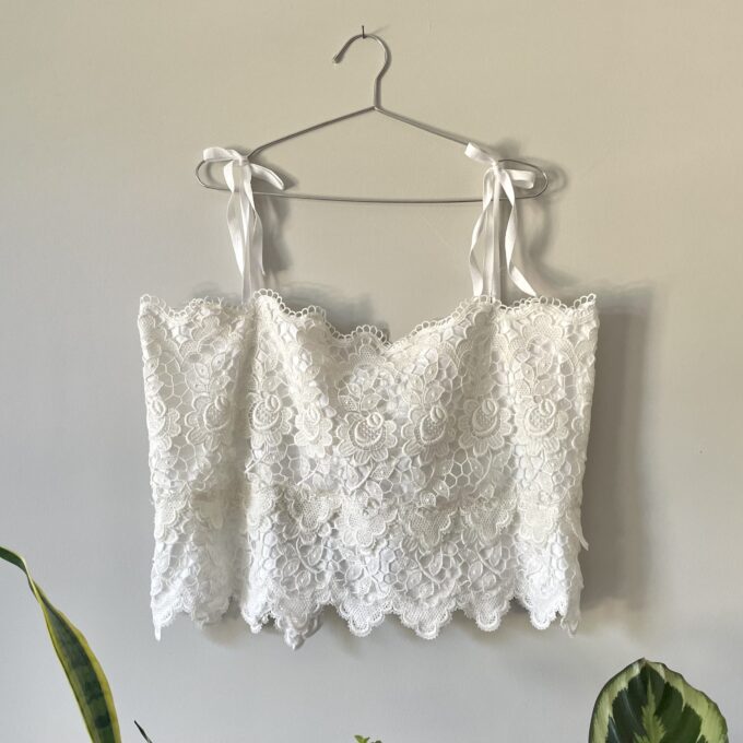 A White Lace Festival Tank Top w/ Tie Back hanging on a wall next to a potted plant.