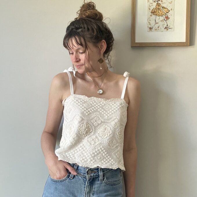 A woman standing next to a wall wearing a cream crochet festival cropped tank top with tie back.
