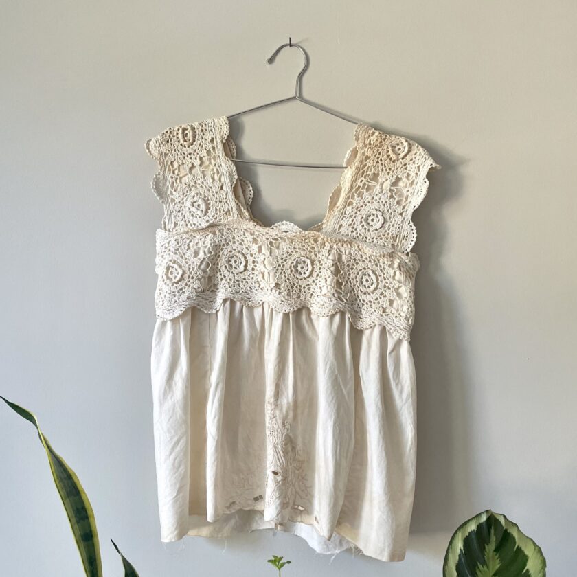 A Tea Stained Cream Crochet Festival Tank Top w/ Tie Back hanging on a wall next to a potted plant.