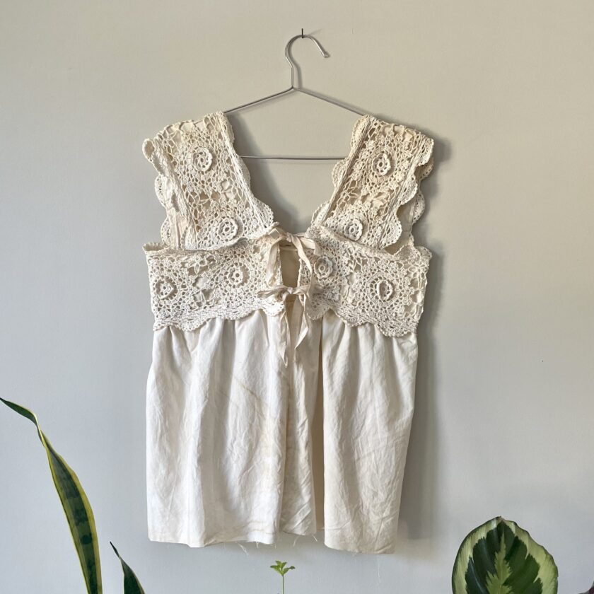 A Tea Stained Cream Crochet Festival Tank Top w/ Tie Back hanging on a wall next to a potted plant.