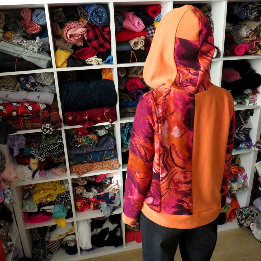 a person standing in front of a Patchwork Marbled Orange Hoodie (Small) hanging in a closet full of clothes.