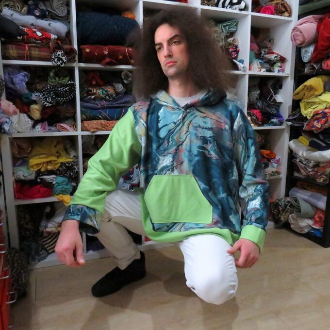 A man with long hair sitting in a closet wearing the Green and Turquoise Marbled Hoodie (3X-Large).