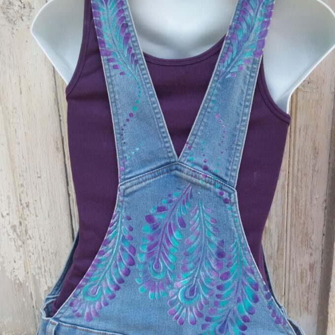 Hand painted ladies denim overalls with distressed details have a lovely purple and teal feather design.
