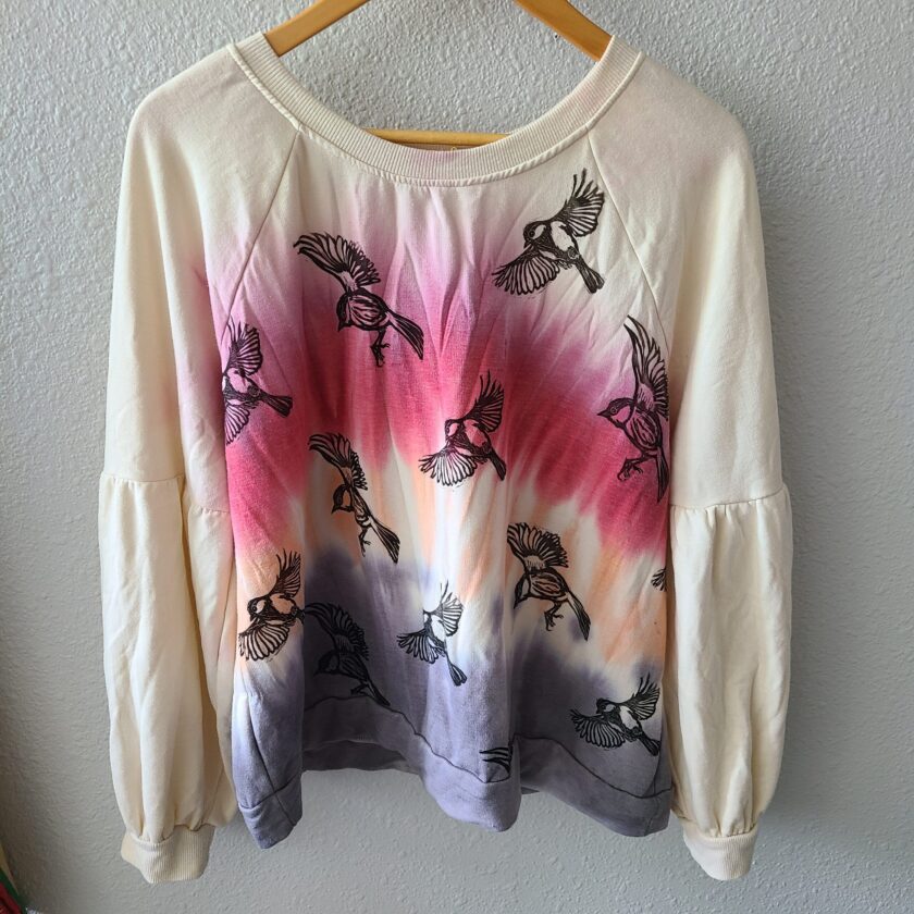 a tie dye sweater with birds on it hanging on a hanger.