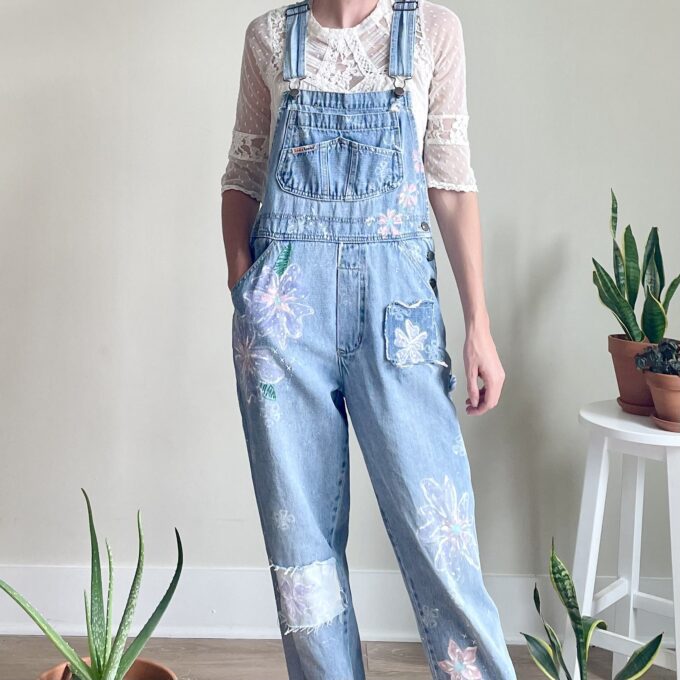 A woman is standing in front of a potted plant wearing denim overalls.