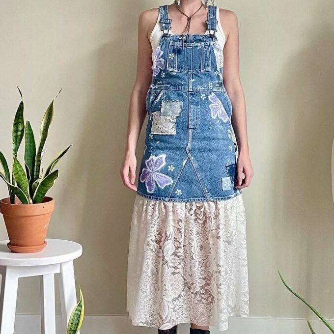 A woman is standing in front of a potted plant wearing a denim overall dress.