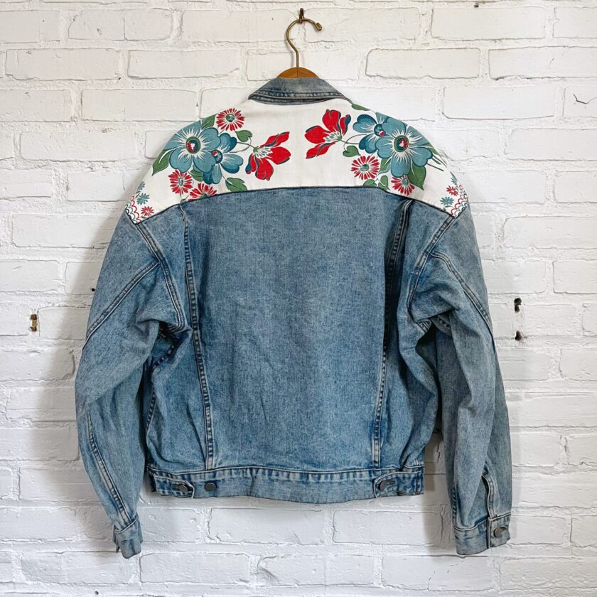 A denim jacket mended with a floral tea towel hanging on a white brick wall.