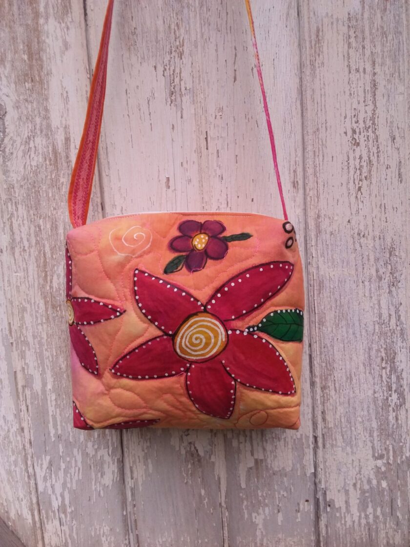 Hand painted red and purple flowers on an orange bag shown hanging on a wall