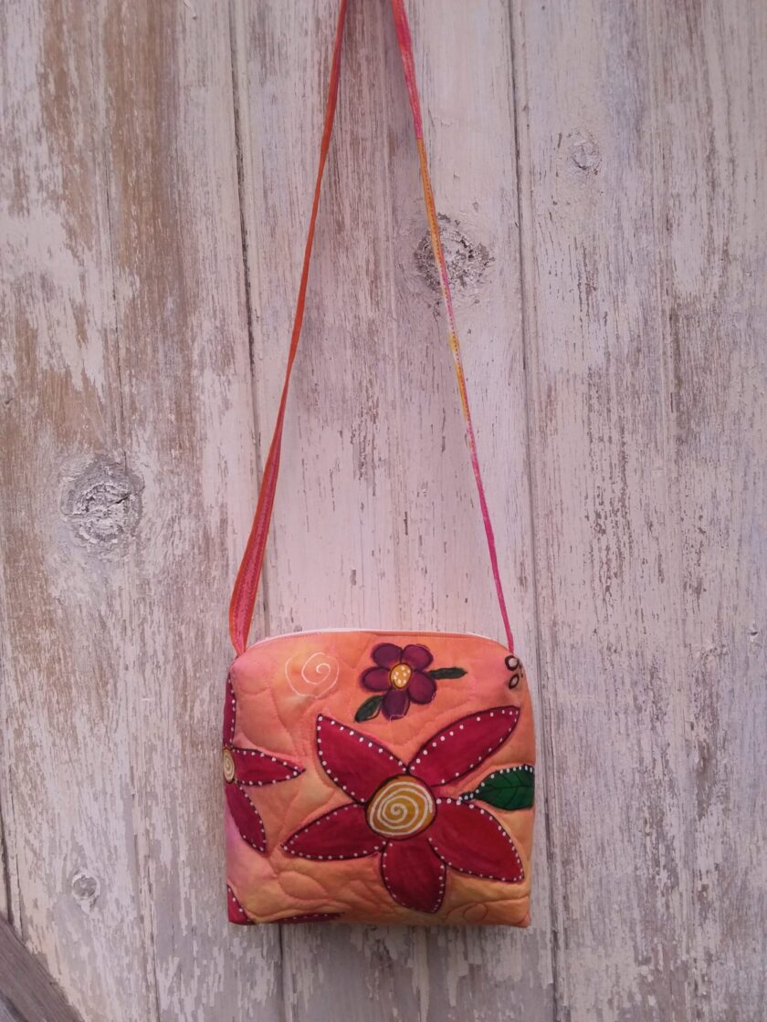A hand painted bag with red and purple flowers and a bright orange upcycled fabric hanging on a wall